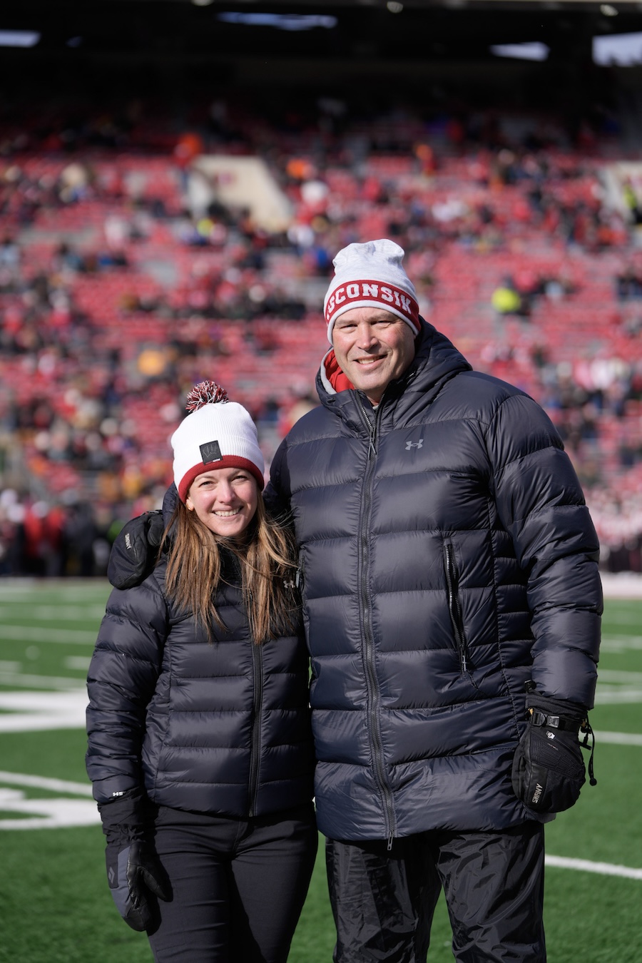 A man and a woman wearing winter coats and Wisconsin labeled hats standing on the Wisconsin Badgers football field.